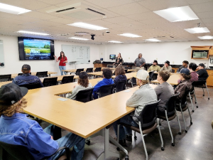 13 Students sit in a classroom watching as a woman gives a presentation.