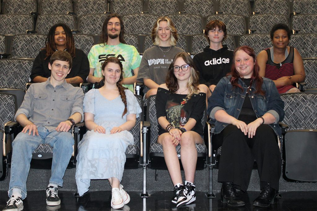 Nine students of different races and genders sit in two staggered rows in a theater.