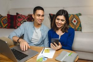 A man and woman are sitting on their floor in front of a table. One has a laptop and the other is holding a phone.