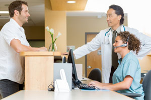 A doctor stands next to a woman in front of a computer screen, both of whom are talking to a patient on the other side of the desk.