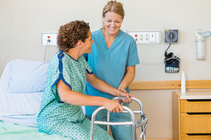 A nurse helps a patient with a walker stand up from the hospital bed.